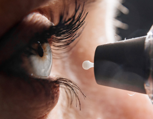 Nahaufnahme eines Auges mit langen Wimpern, vor dem eine Pipette mit einem Tropfen Flüssigkeit zu sehen ist.