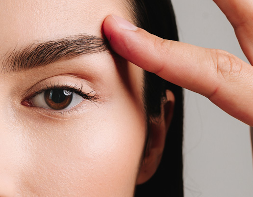 Hand berührt die Stirn einer Person, die Augenbraue ist sichtbar, Hintergrund grau.